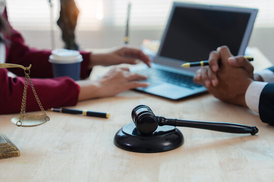 Close up of justice scales and judge gavel on wooden desk with cash bundle and lawyer consulting client about financial lawsuit in blurred background.