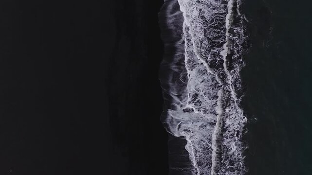 Aerial top down view of Vik, Iceland black sand beach as waves roll in parallel lines and break into lace like white surf, shot in overcast daylight with steady motion