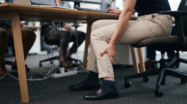 Closeup of a woman sitting at an office desk, holding her leg and experiencing calf pain, suggesting discomfort or a medical issue
