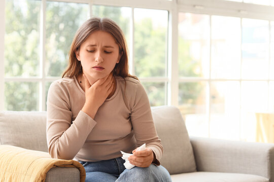Young woman suffering from sore throat and holding a tissue while sitting on a sofa at home, She holds a tissue, indicating symptoms of illness such as a cold or flu
