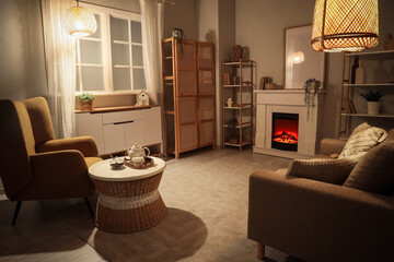 Obraz premium Cozy living room interior with warm lighting, comfortable armchairs, and a decorative electric fireplace at night