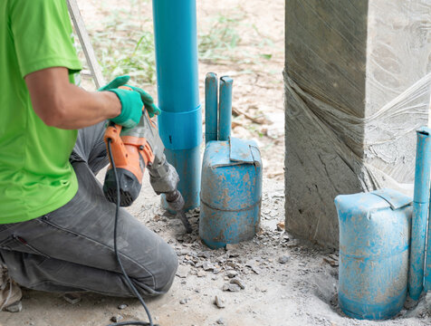 Construction worker using an electric jackhammer to break up concrete around blue plumbing utility pipes.