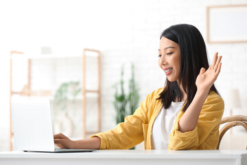 Beautiful young Asian woman with laptop waving hand at home © Pixel-Shot