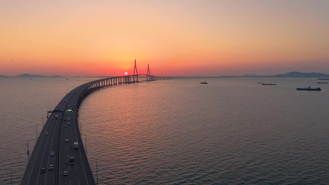 Aerial view shows sun setting directly behind Incheon Bridge cable tower as traffic crosses curving causeway over calm sea
