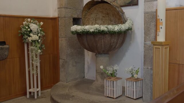 Round baptism font wreathed in white flowers beside paschal candle indoors
