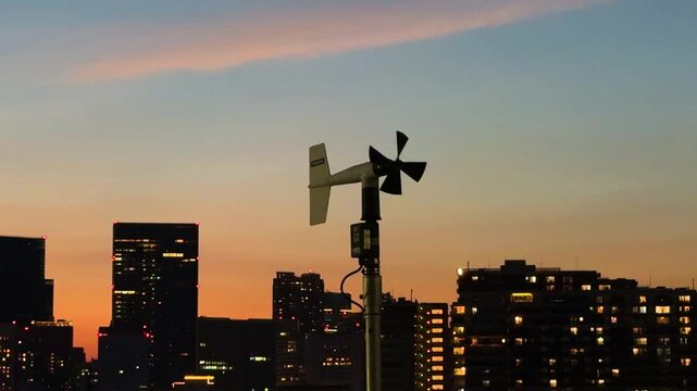 夕焼け空と都市ビル群の夜景を背景にたたずむ風向風速計のシルエットと夕暮れの気象観測風景。