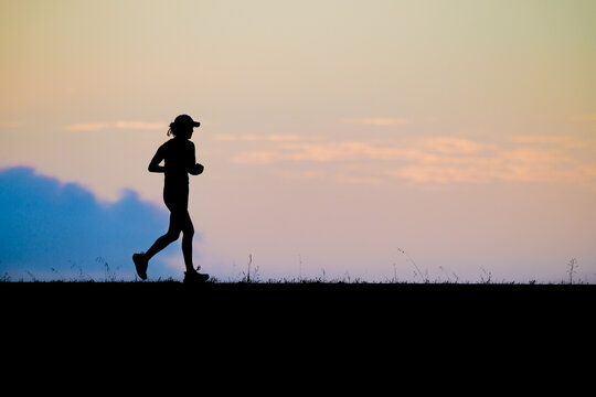Side view silhouette of a female runner jogging at sunset along a scenic horizon with warm orange tones and soft blue sky showcasing fitness endurance active lifestyle and motivation.