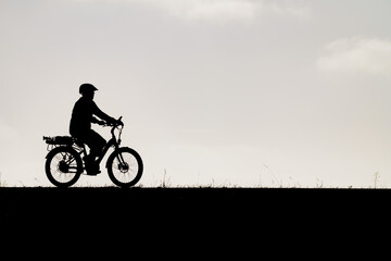 Obraz premium Dark silhouette of a cyclist riding an electric bike with a rear cargo rack along a smooth horizon under a bright sky during an outdoor recreational bicycle outing.