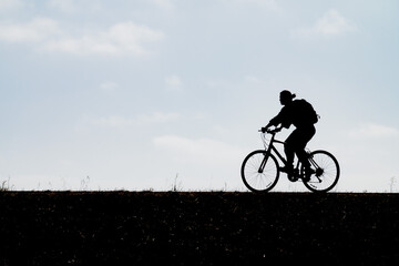 Obraz premium Dynamic silhouette of a cyclist wearing a backpack riding a bicycle across a high ridge line against a bright afternoon sky emphasizing active commuting healthy lifestyle and outdoors exercise.