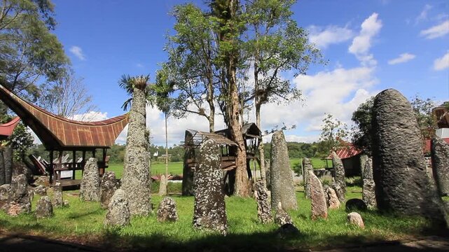 Ancient megalithic stone pillars (menhirs) standing in Bori Kalimpenne burial site under bright sunny sky with traditional Toraja Tongkonan house and lush green surroundings, South Sulawesi, Indonesia