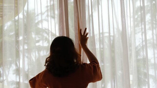 The woman was at the window in the bedroom. She opened the curtains on the window. In the morning and she looks at the view of mountains and trees at sunrise.	