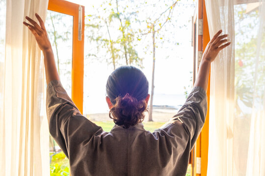 The woman was at the window in the bedroom. She opened the curtains on the window. In the morning and she looks at the view of mountains and trees at sunrise.	