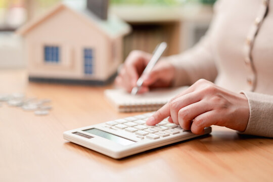 Mortgage calculation and home budgeting concept. Woman using calculator and writing on notebook with blurred house model.