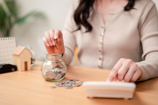 Personal finance and saving goals concept. Woman hand dropping a coin into a glass jar with house and car models on desk.