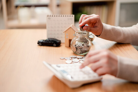Personal finance and saving goals concept. Woman hand dropping a coin into a glass jar with house and car models on desk.