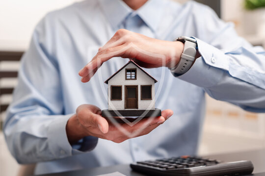 Property insurance and home protection concept. Businessman hands protecting a model house with shield icon.