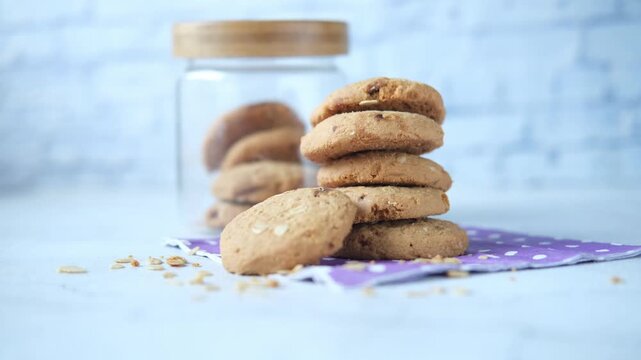 Stack of fresh homemade oatmeal cookies on purple napkin with jar of biscuits in background on white table