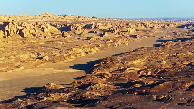 Aerial view of vast desert yardang landform with rocky cliffs, sandy terrain, and dramatic sunlight.