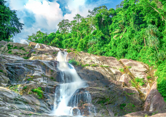 Beautiful tropical waterfall cascading over granite mountain rocks beneath blue sky. Lush rainforest in Thailand. Wilderness landscape for travel and nature theme. Ecotourism and conservation. © Fahroni