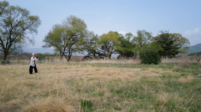 Lone figure walks through open grassland beneath a clear sky. Trees stand in rows, green and bare against the distant hills. The person moves slowly, absorbed in quiet exploration