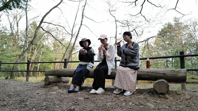 Three friends sit on a rustic wooden bench. They enjoy a calm moment in a quiet park setting. Bare trees frame the background softly. Their relaxed poses suggest friendship and contentment