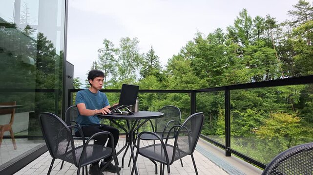 Man sits calmly on balcony, typing on laptop. Outdoor table, chairs surround him. Garden view, trees gently sway behind glass. He appears focused, calm, content with work. Ideal for modern lifestyle