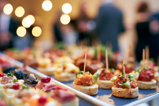 Delicious appetizers and canap&eacute;s laid out on a table at a party, with blurred figures of people in the background. The food looks colorful and appetizing, ready to be enjoyed.