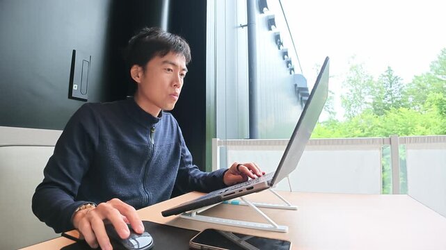 Young man focuses on laptop work. He uses mouse, sits upright, eyes fixed forward. Natural light streams through wide window behind him. Modern office space, calm atmosphere