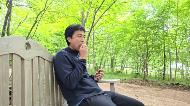 Man sits quietly on park bench. He gazes off into distance with calm expression. Hands gently hold small object near his chest. Green trees frame peaceful, sunlit outdoor setting