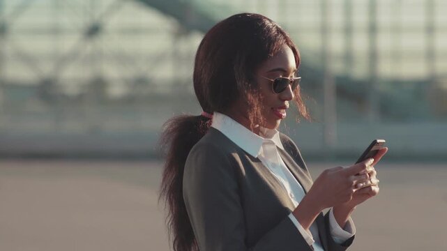 Beautiful long-haired Afro-American woman in trendy sunglasses actively uses her smartphone outside the office, gets good news, looks happy and satisfied. Positive emotions, cheerful mood, feeling