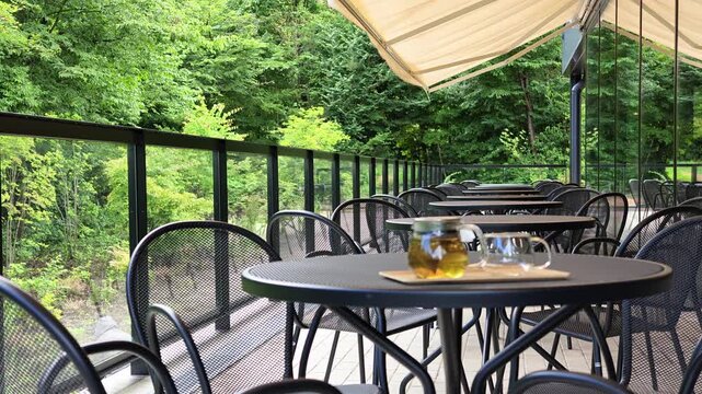 Outdoor cafe table set for tea. Green trees frame the scene. Black metal furniture adds contrast. Cream awning offers gentle shade. Serenity radiates through stillness