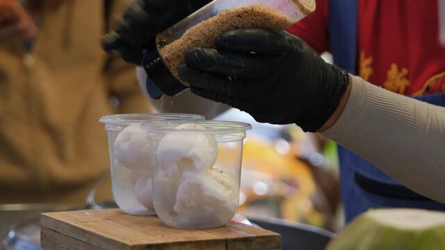 Street vendor drizzling coconut milk and sprinkling crushed nuts over a freshly prepared coconut ice cream serving at an outdoor street market. Thai street food concept
