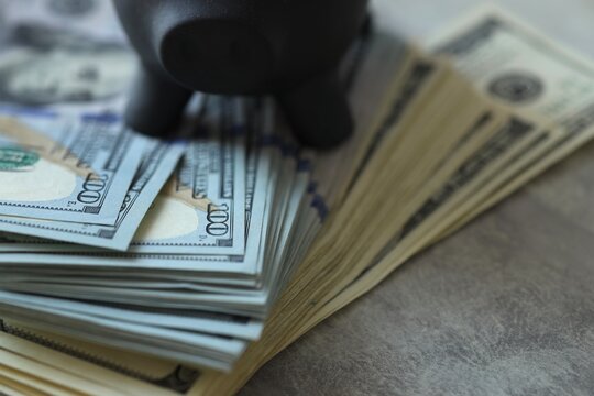 A piggy bank is placed on top of a large amount of cash. The bills are arranged in stacks and show various values. The scene is indoors on a simple surface.