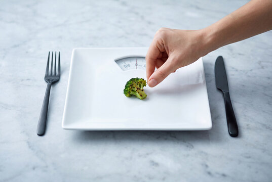 Hand placing single broccoli floret on empty plate resembling scale