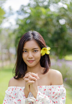 Portrait of a young Asian woman with a yellow flower behind her ear, clasping her hands together in a gentle and polite gesture, outdoors in a green park with soft natural lighting