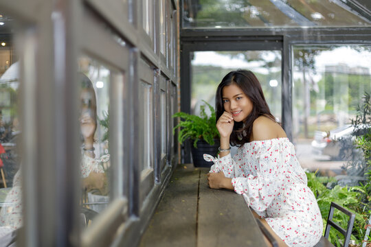 Portrait of a smiling young Asian woman sitting at a wooden counter inside a cozy cafe, leaning on her arms with a reflection in the window glass, surrounded by green plants.