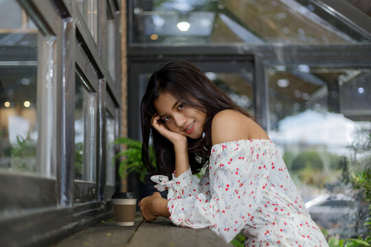 Portrait of a smiling young Asian woman resting her head on her hand, sitting at a wooden counter with a takeaway coffee cup in a cozy cafe with glass windows and greenery.