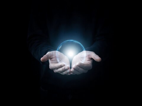 Human hands cupped together holding a glowing blue sphere of light against black background