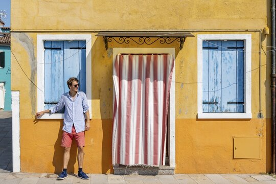 Young man leaning against house wall, yellow house facade with entrance door with red and white curtain and windows with blue shutters, colourful houses on the island of Burano, Venice, Veneto, Italy