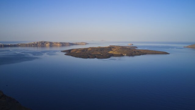 Drone shot, morning atmosphere, caldera, view of volcanic islands Nea Kameni, Palea Kameni and Aspronisi, view of southern tip of the island, sea smooth as glass and blue, sky blue and cloudless, caldera in front of Firostefani, Santorini Island, Cyclades, Greece