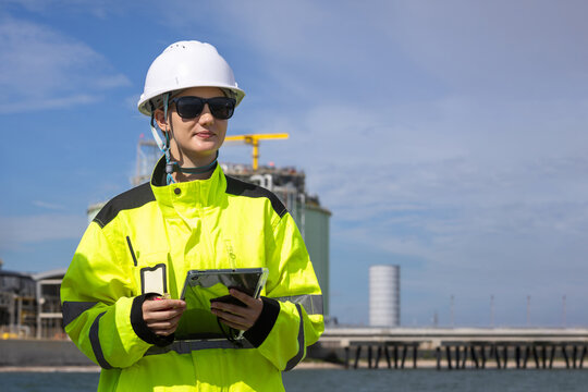 Female industrial engineer using tablet at LNG plant facility, Technician checking data on device at natural gas terminal, Woman supervisor in safety gear managing operation at industrial storage site