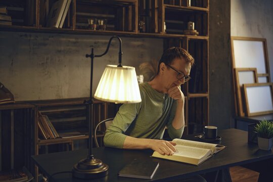 A man sits at a desk in a dimly lit room, engrossed in a book. Warm light from a lamp highlights the pages while a cup rests by his side. Bookshelves filled with books create a calming atmosphere