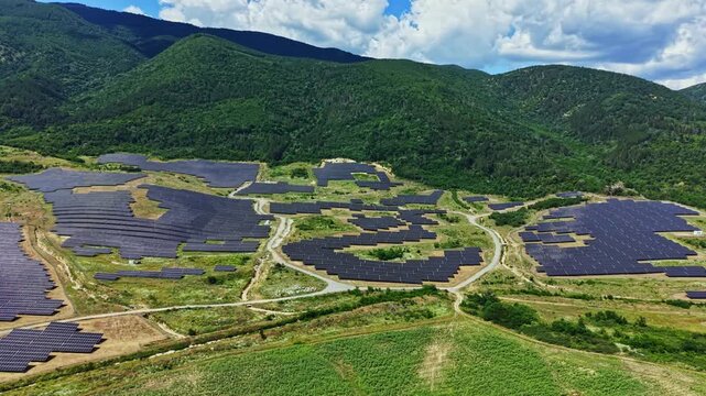 Rows of solar panels stretch across fields in a rural area. Green hills and mountains are visible in the background. Clear sky with few clouds is seen above.