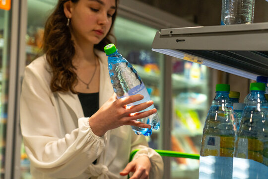 A Young Woman Examining a Bottle of Water in a Store Aisle Amidst Shelves of Various Products While Shopping for Refreshments.
