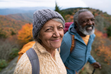 Fototapeta premium Older couple smiles warmly on autumn trail. Backpacks hint at a scenic hike. Mountain range stretches behind them. Vibrant fall colors surround the path. Their joy reflects shared adventure