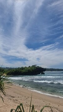 Sandy tropical beach with gentle waves and a green rocky island under a wide cloudy blue sky