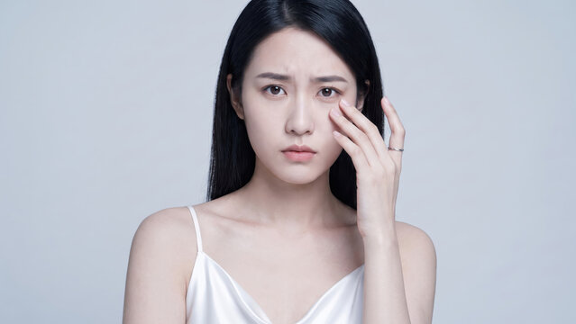 Young Asian woman with a concerned expression gently touching her under-eye, looking at the camera for beauty and skincare.