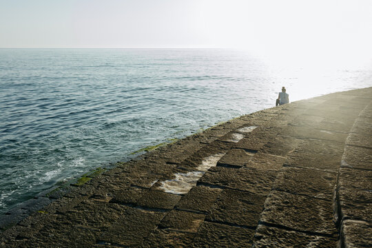 Back view of a person sitting on a stone pier by the sea