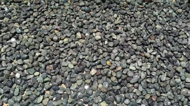 water gently spraying over a bed of wet, dark pebble stones and black river rocks, creating a glistening, reflective texture. The steady stream of water droplets highlights the natural, smooth shapes 