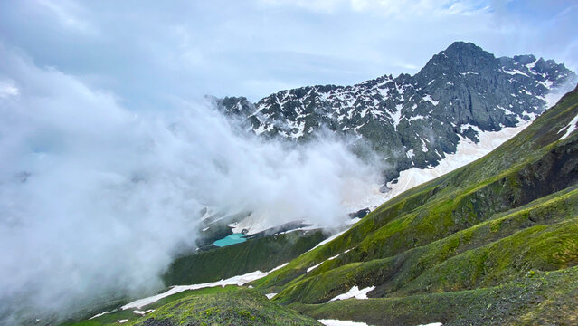Clouds reveal White Abudelauri Lake below Chaukhi Pass in Georgia, where turquoise glacial water, snowfields and steep slopes